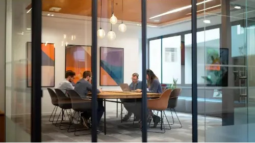 A group of people are sitting around a table in a conference room. The room is well lit and has a modern design. The people are working on a project and are using laptops